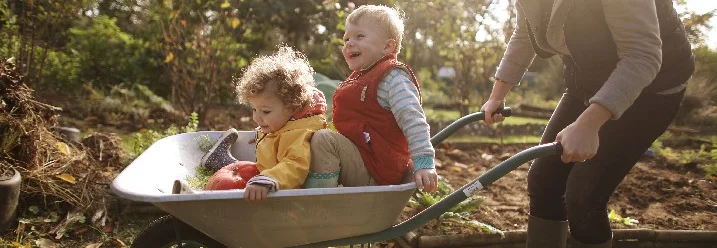 Zwei Kinder in Schubkarre, Person schiebt.